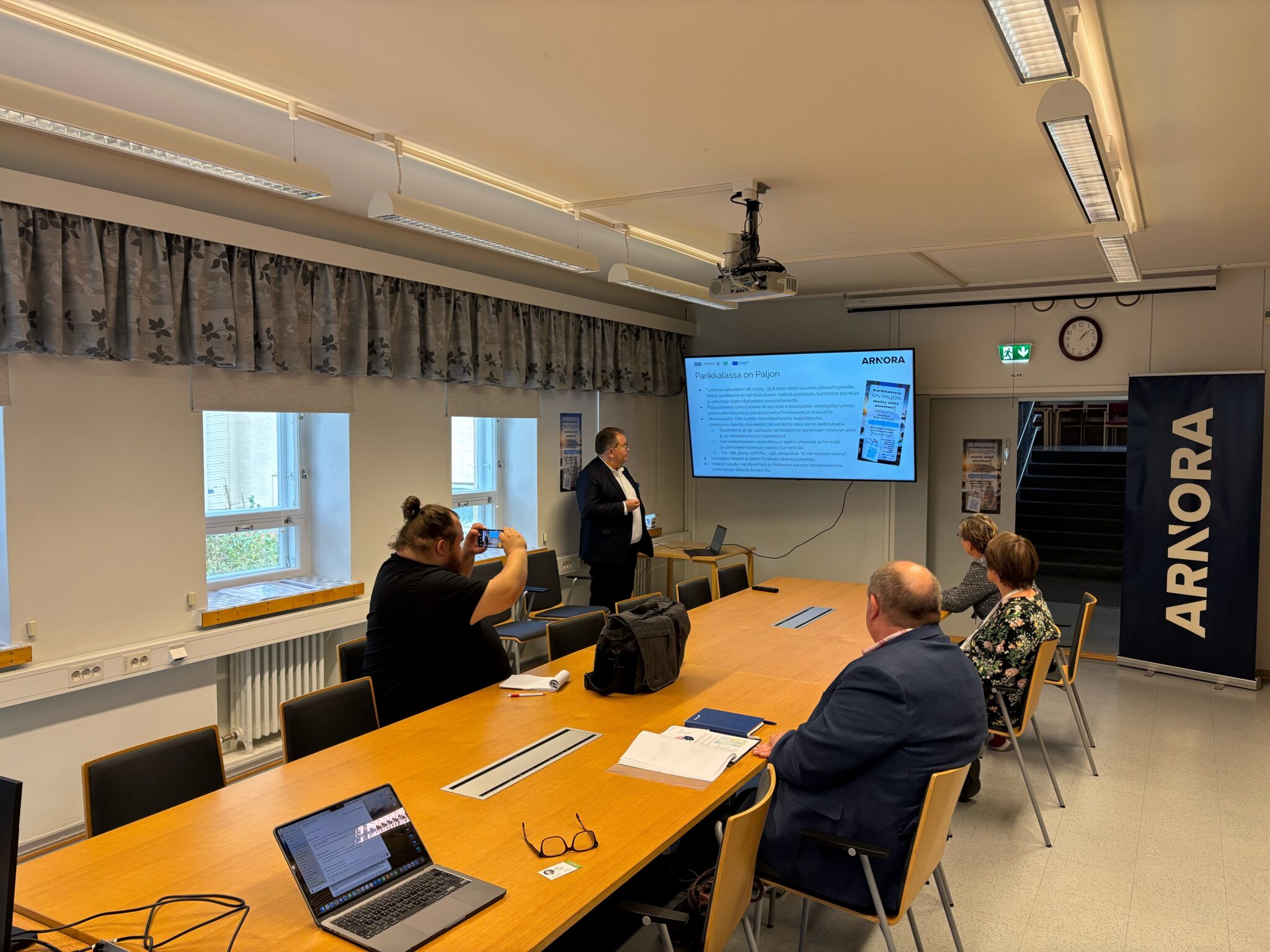 Kokouspöydän ääressä kolme henkilöä seuraamassa esittelijän presentaatiota.