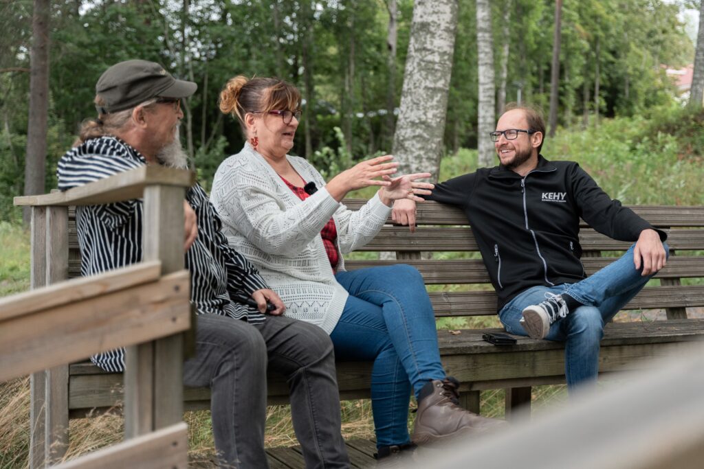 Nainen ja kaksi miestä istuvat kesällä penkillä.
