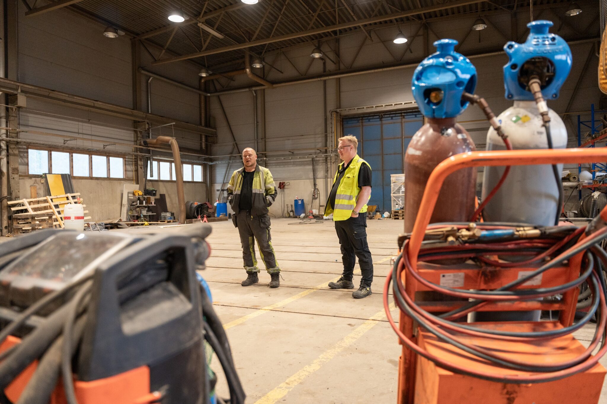 Konehallissa hitsauskalustoa, taustalla kaksi henkilöä työvaatteissa.