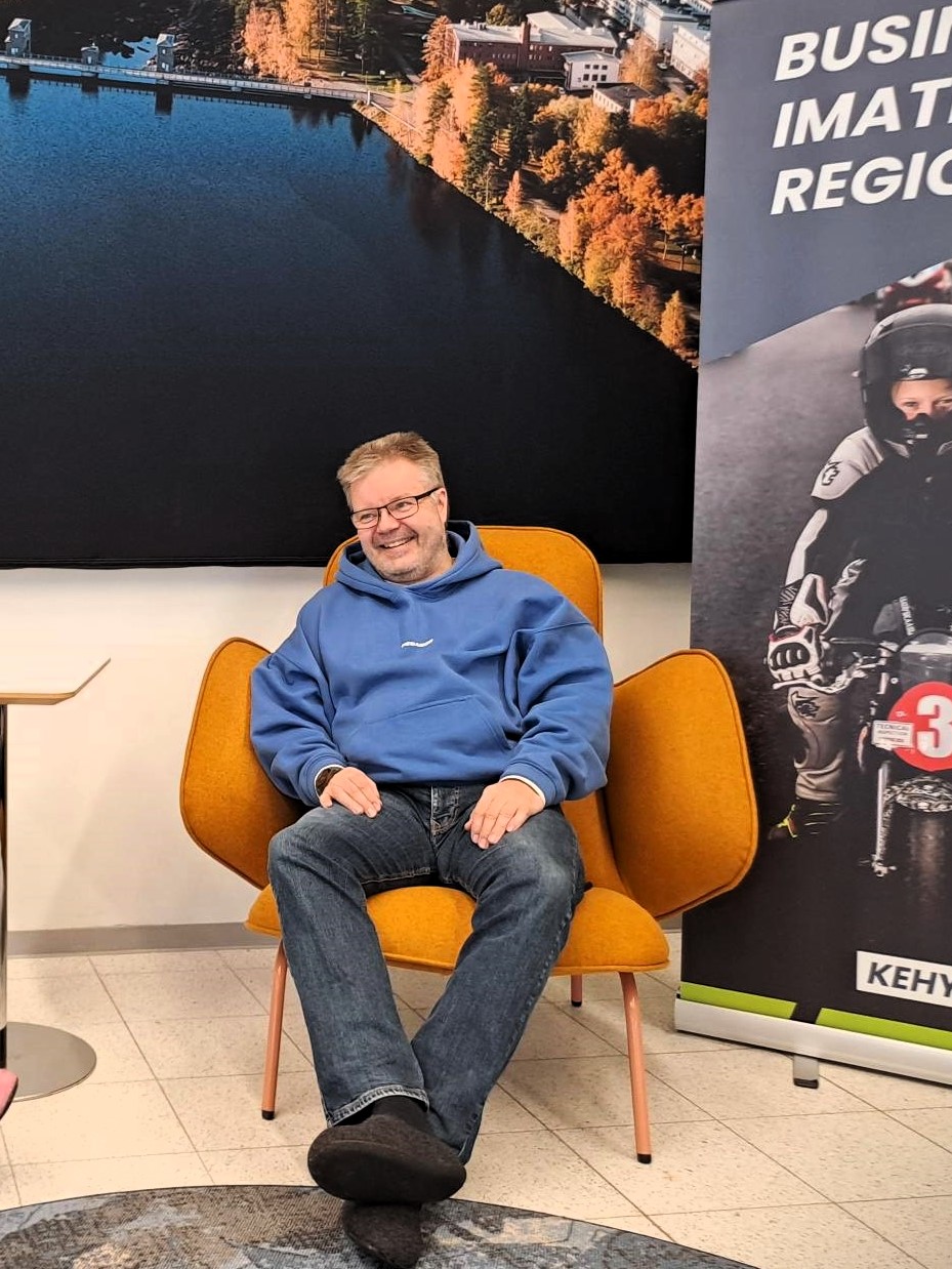 Mies istuu oranssissa nojatuolissa, taustalla seinäkuva Vuoksesta ja Business Imatra Regionin roll up-mainos.